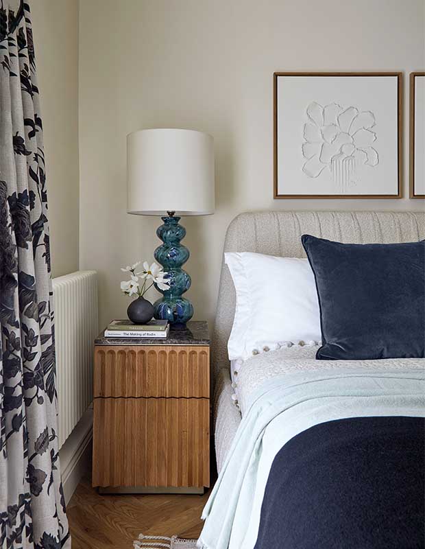 A blue and white principal bedroom is pretty and tied together. Navy blue elements such as pillows, blankets, and patterned drapes with white walls and background details are tied together with, light wood accents. notting hill flat